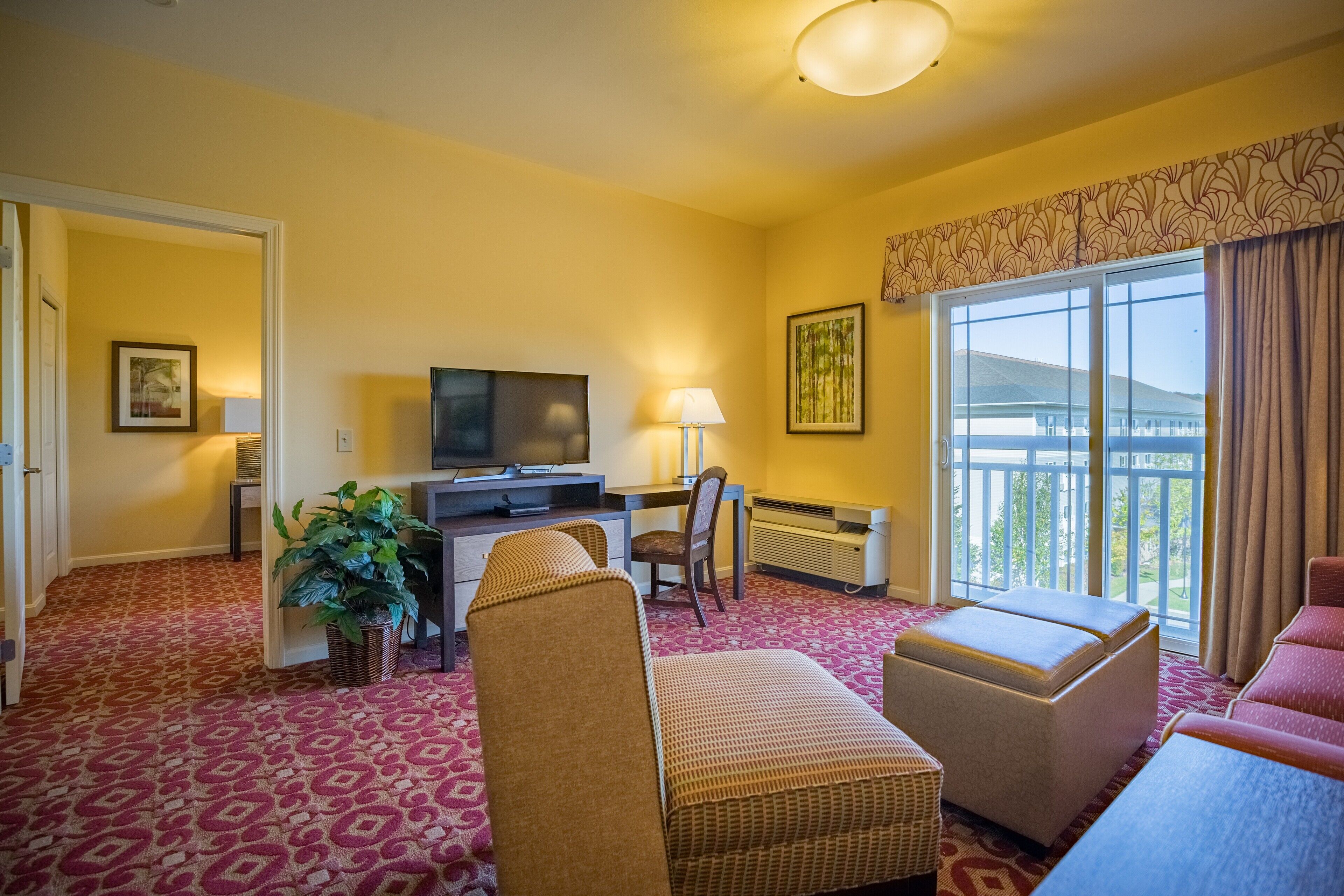 A hotel room with a large window, a desk with a computer, a TV, a chair, a sofa, and a potted plant.
