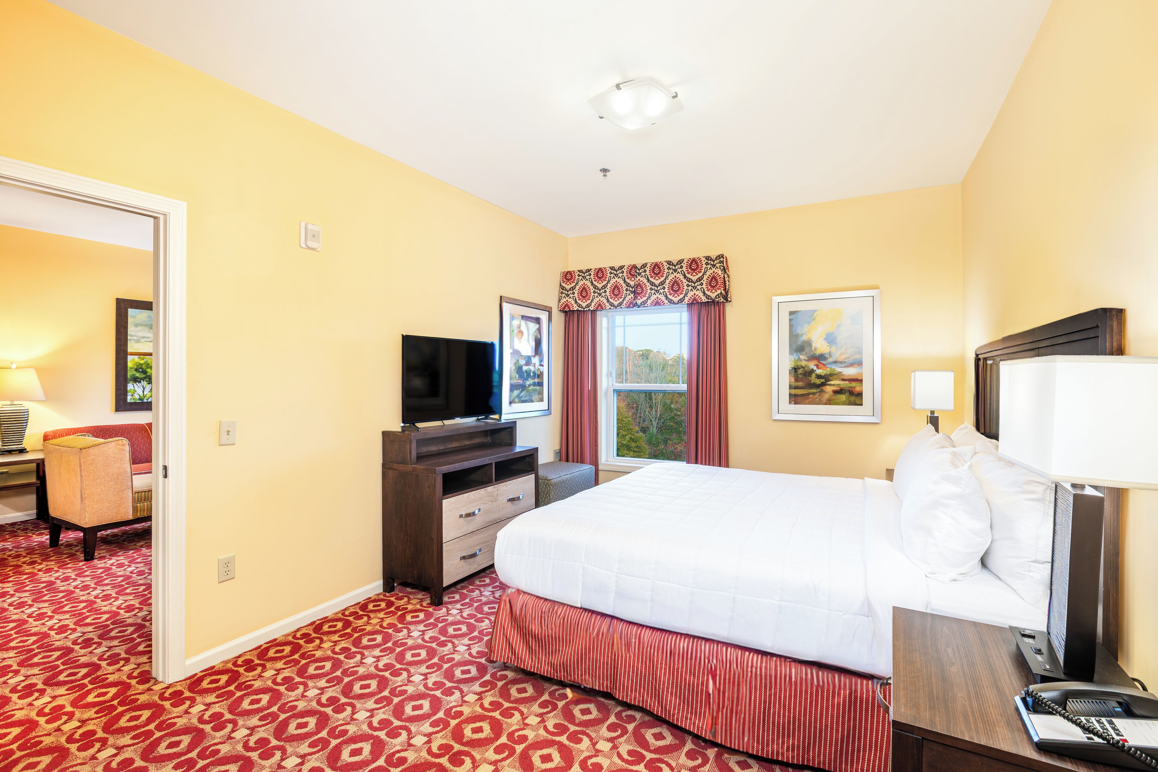A hotel room with a large bed, a TV, a desk with a lamp, and a window with curtains.