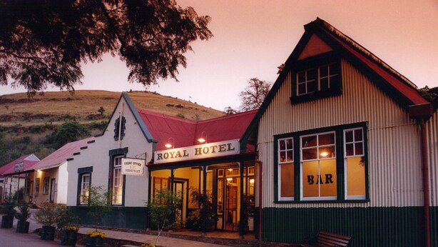 A hotel with a sign reading "ROYAL HOTEL" and a bar sign. There are potted plants and a bench outside. - The Royal Hotel (Pilgrims Rest)