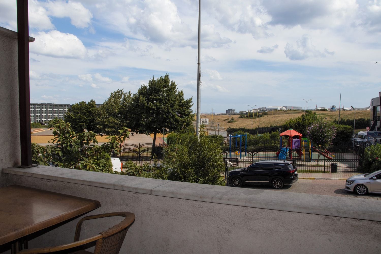 A balcony with a view of a playground, parked cars, and a building in the background.