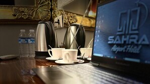 A laptop on a wooden desk with a coffee setup, a water bottle, and a kettle. The laptop screen displays "LID SAHRA Airport Hotel". - Sahra Airport Hotel (Istanbul)
