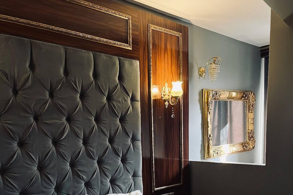 Une chambre d’hôtel avec une tête de lit capitonnée, un grand miroir doté d’un cadre orné et un lit recouvert d’une courtepointe blanche.