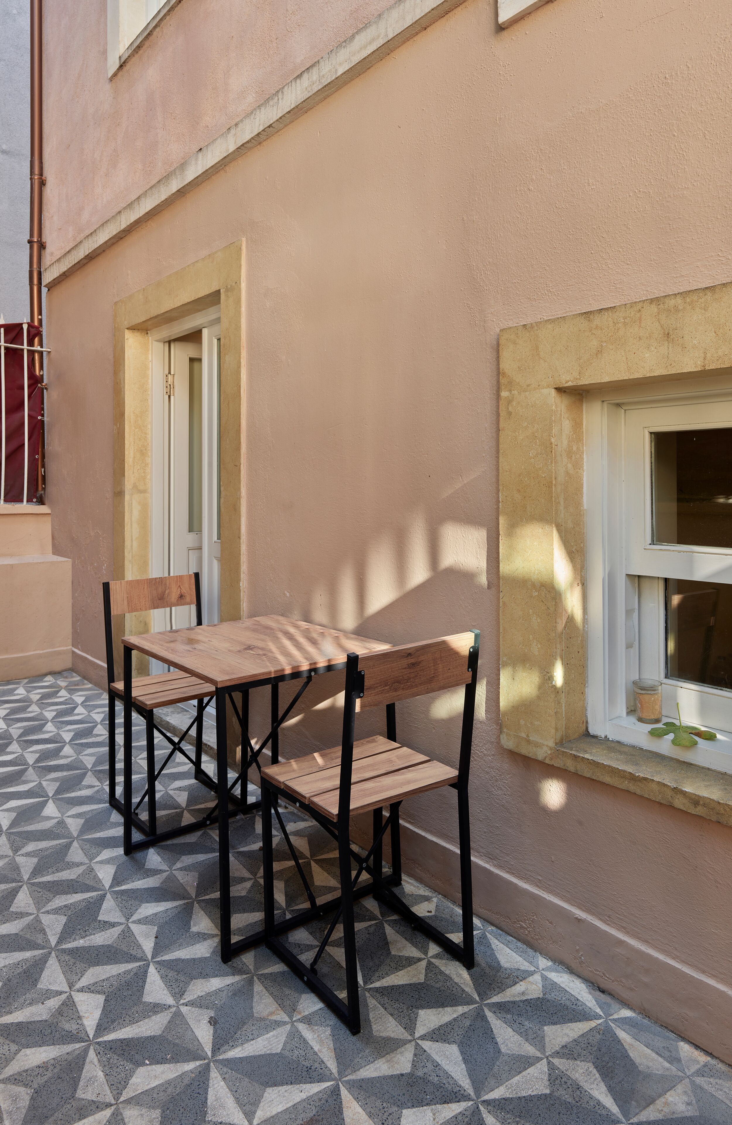 Um pátio com mesa e cadeiras de madeira, chão de azulejos e um edifício com fachada bege e janelas.