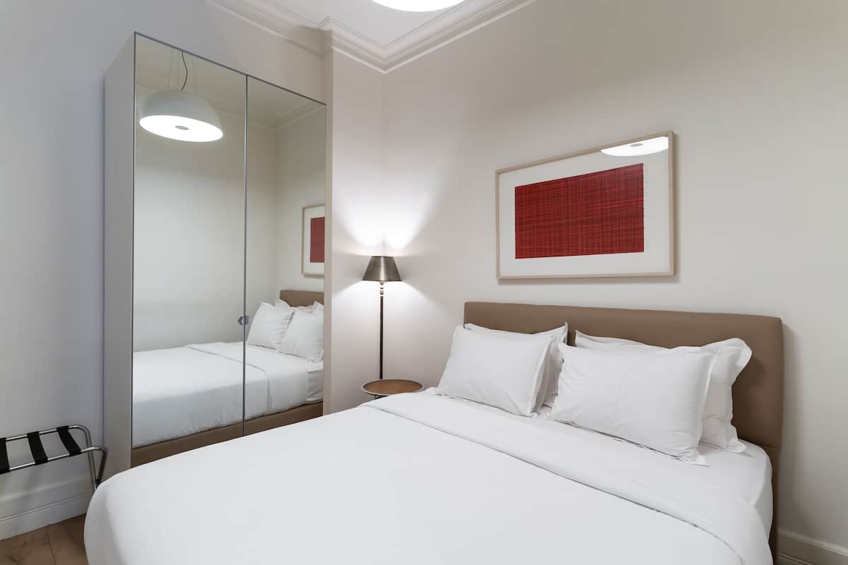A modern hotel room with a large bed, a bedside table, a lamp, a mirror, and a framed artwork on the wall.