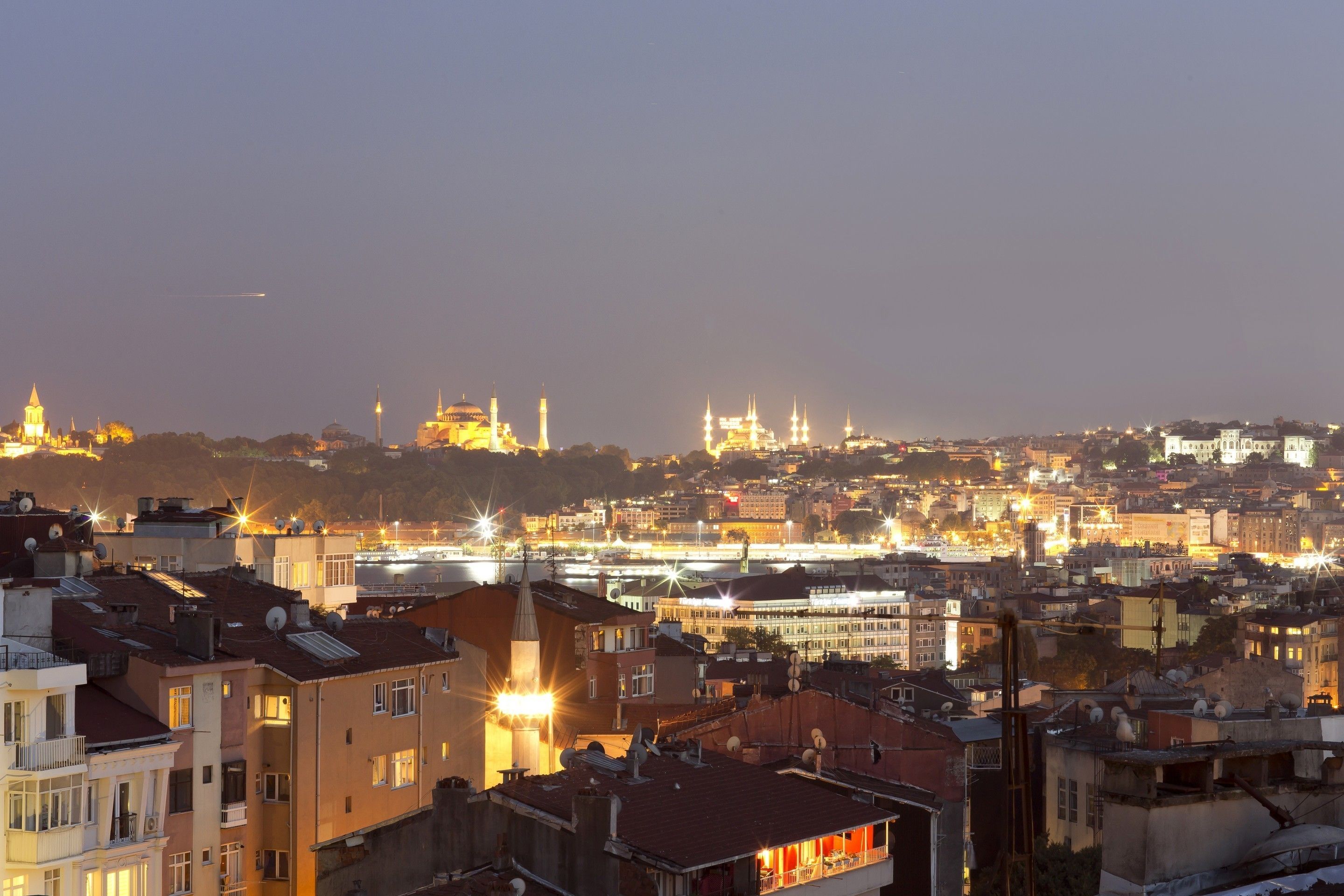Paisagem urbana ao entardecer, com edifícios iluminados e uma mesquita imponente com várias minaretes ao fundo.