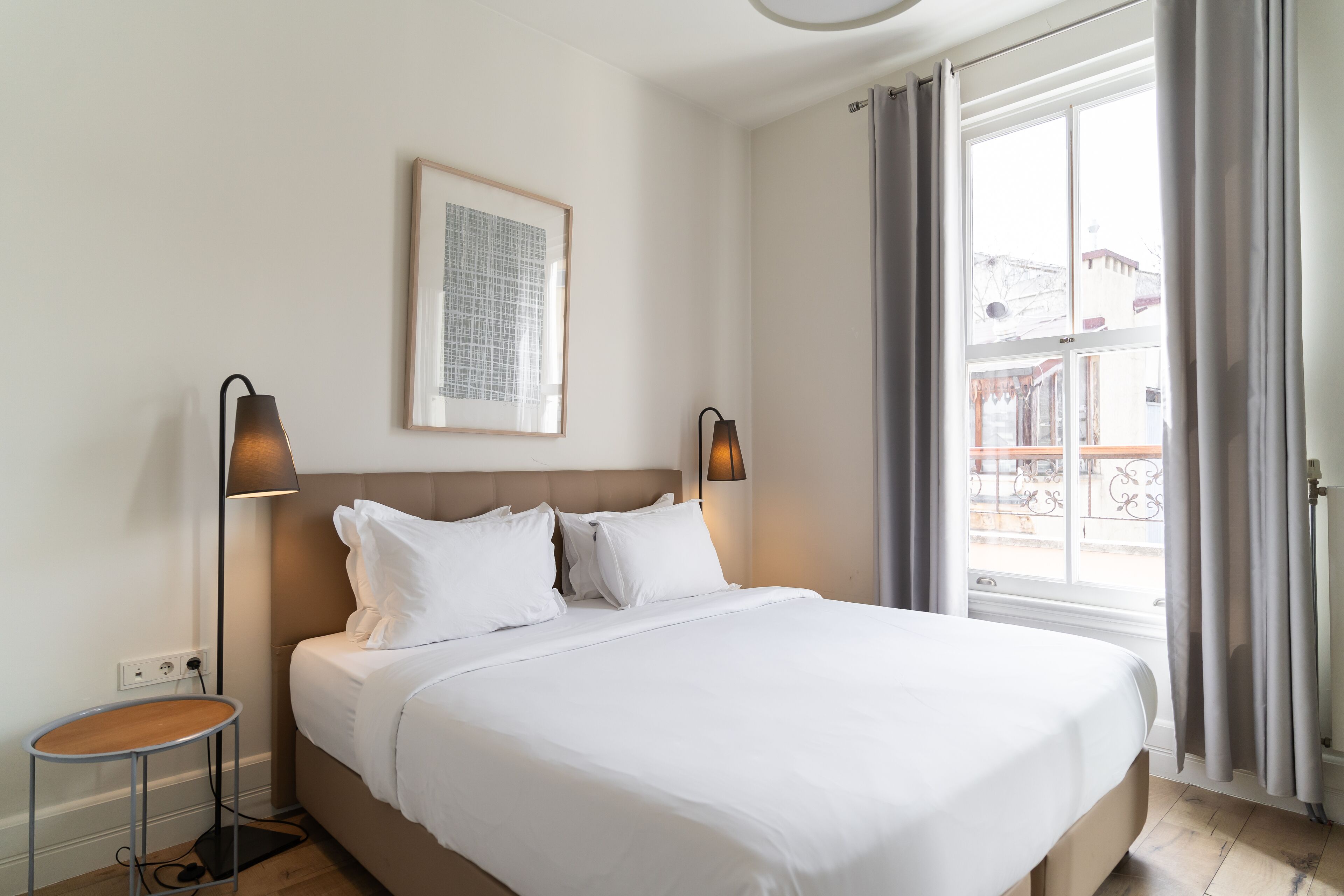 A neatly made bed in a hotel room with a large window, two bedside lamps, and a framed picture on the wall.