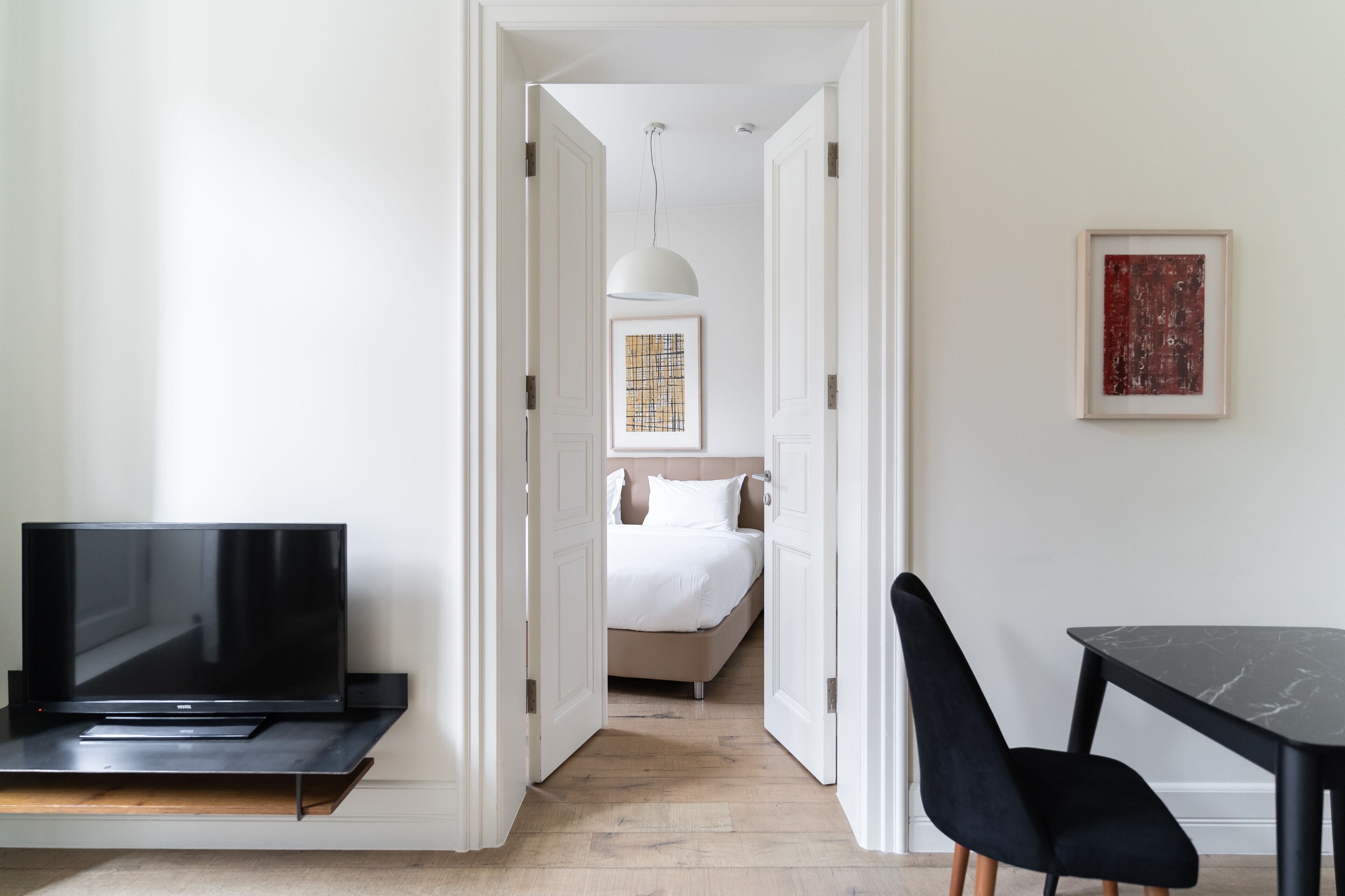 Ein Hotelzimmer mit Fernseher auf einem Schrank, einem Bett mit weißer Bettwäsche, einem Esstisch mit Stuhl und einem gerahmten Kunstwerk an der Wand.