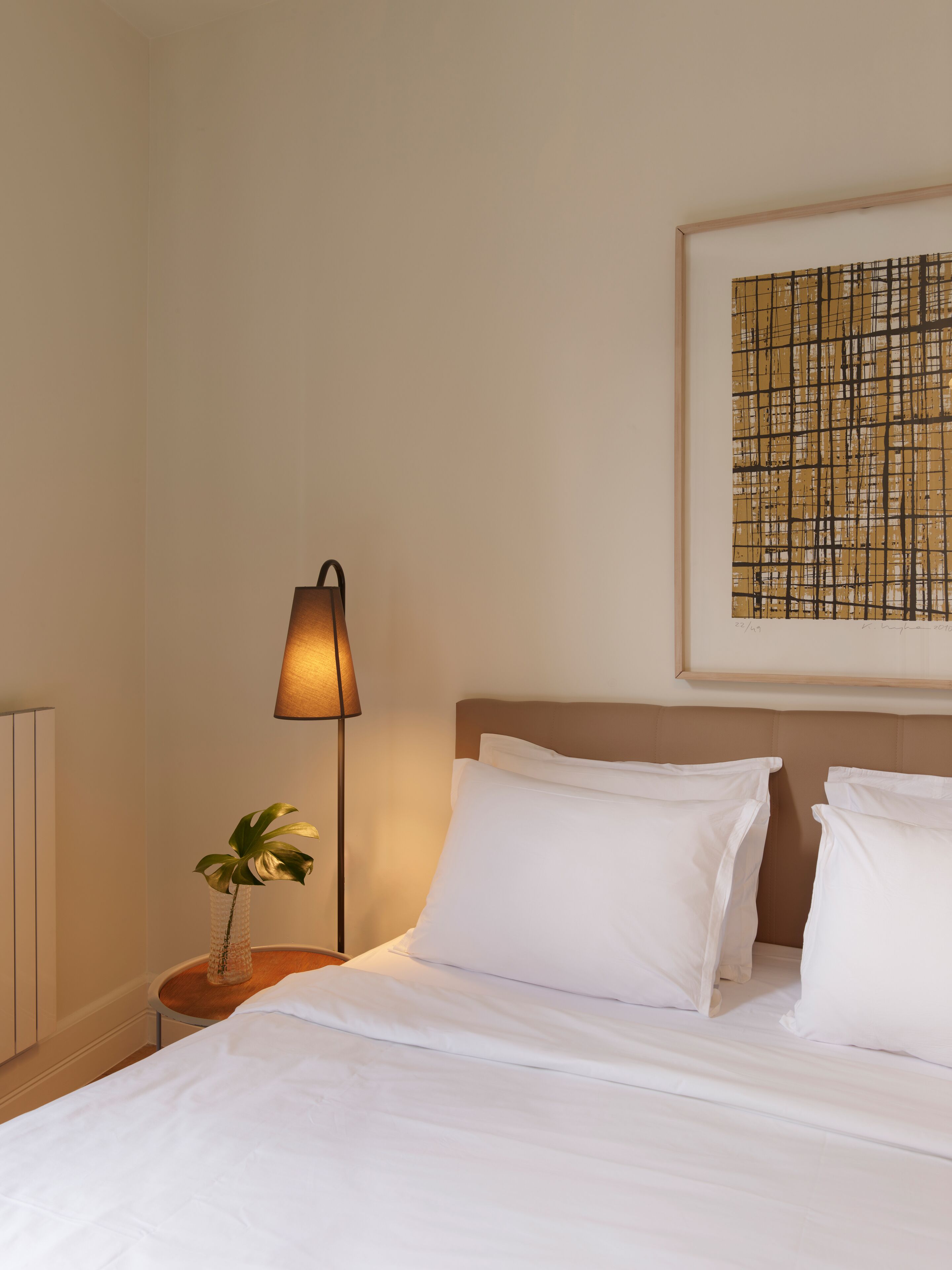 Een net opgemaakt bed met witte lakens, een bedlamp, een kleine potplant en een lijst met abstracte kunst aan de muur.