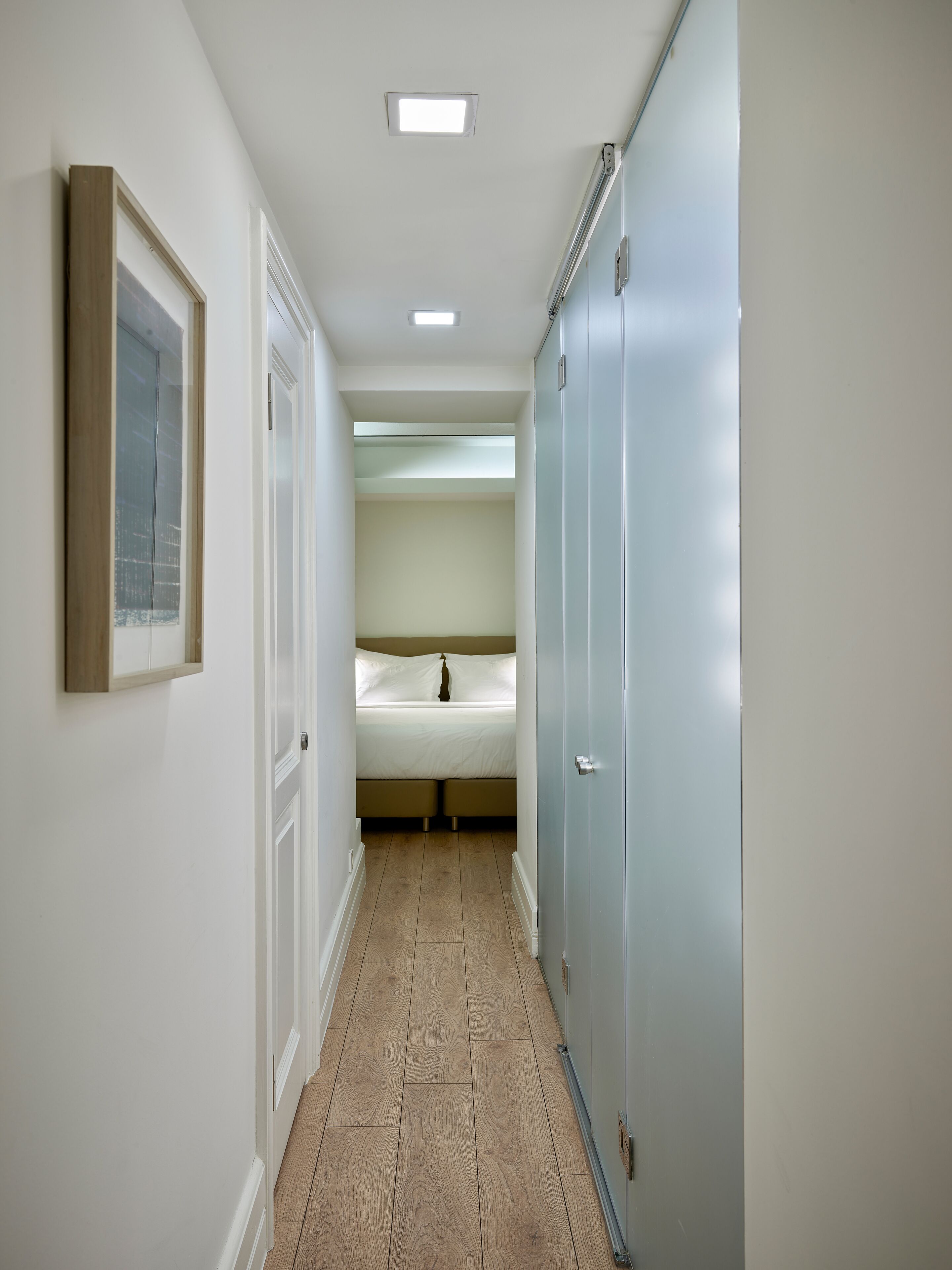 Un pasillo estrecho con suelo de madera que conduce a un dormitorio con cama y armario.