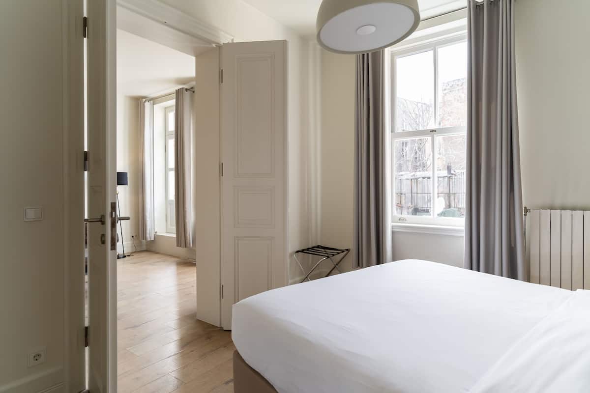 A hotel room with a bed, a radiator, a window with curtains, and a door leading to another room.