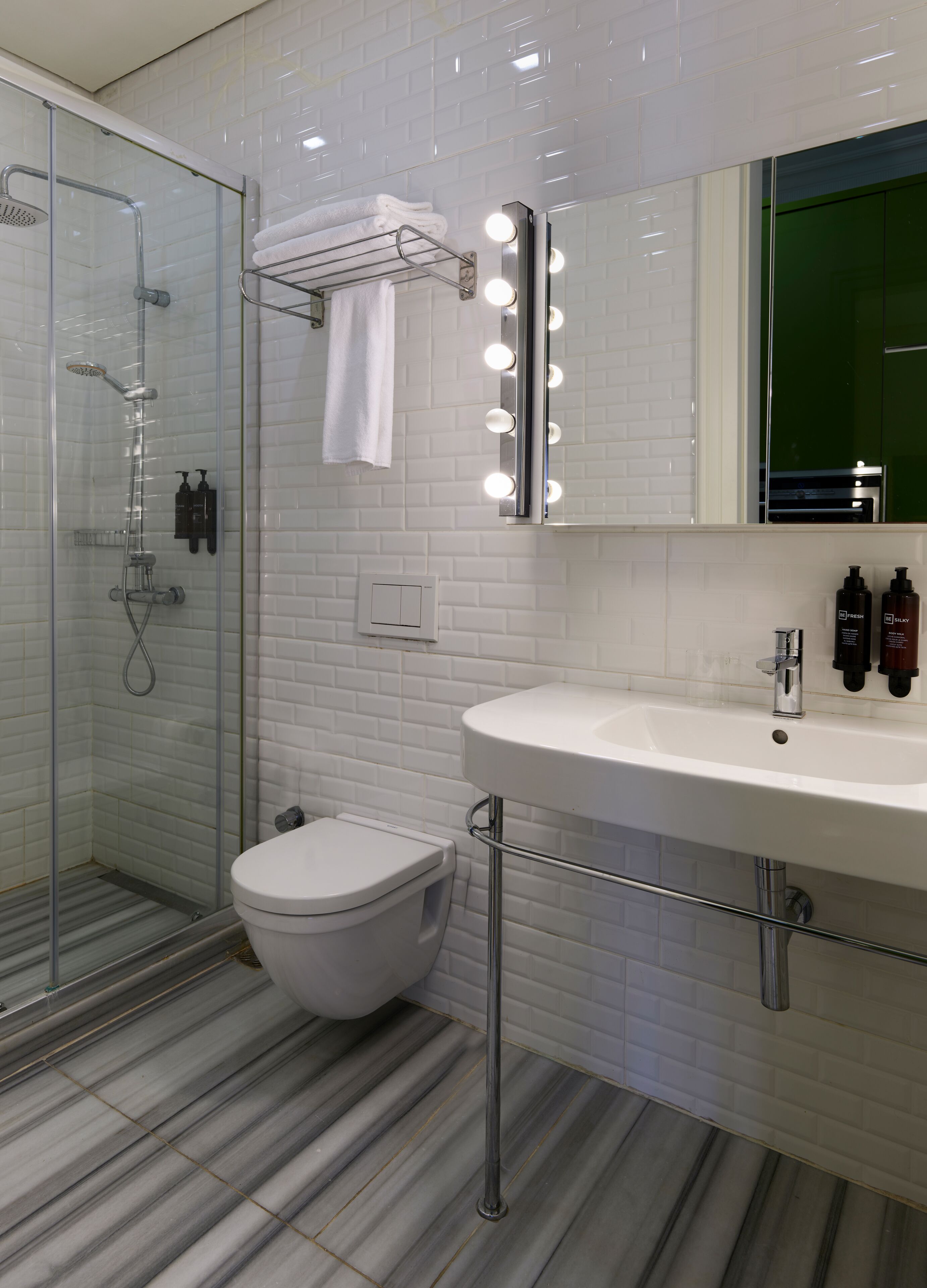 A modern bathroom with a glass shower enclosure, a white toilet, a vanity with a large mirror and lights, and a striped floor.