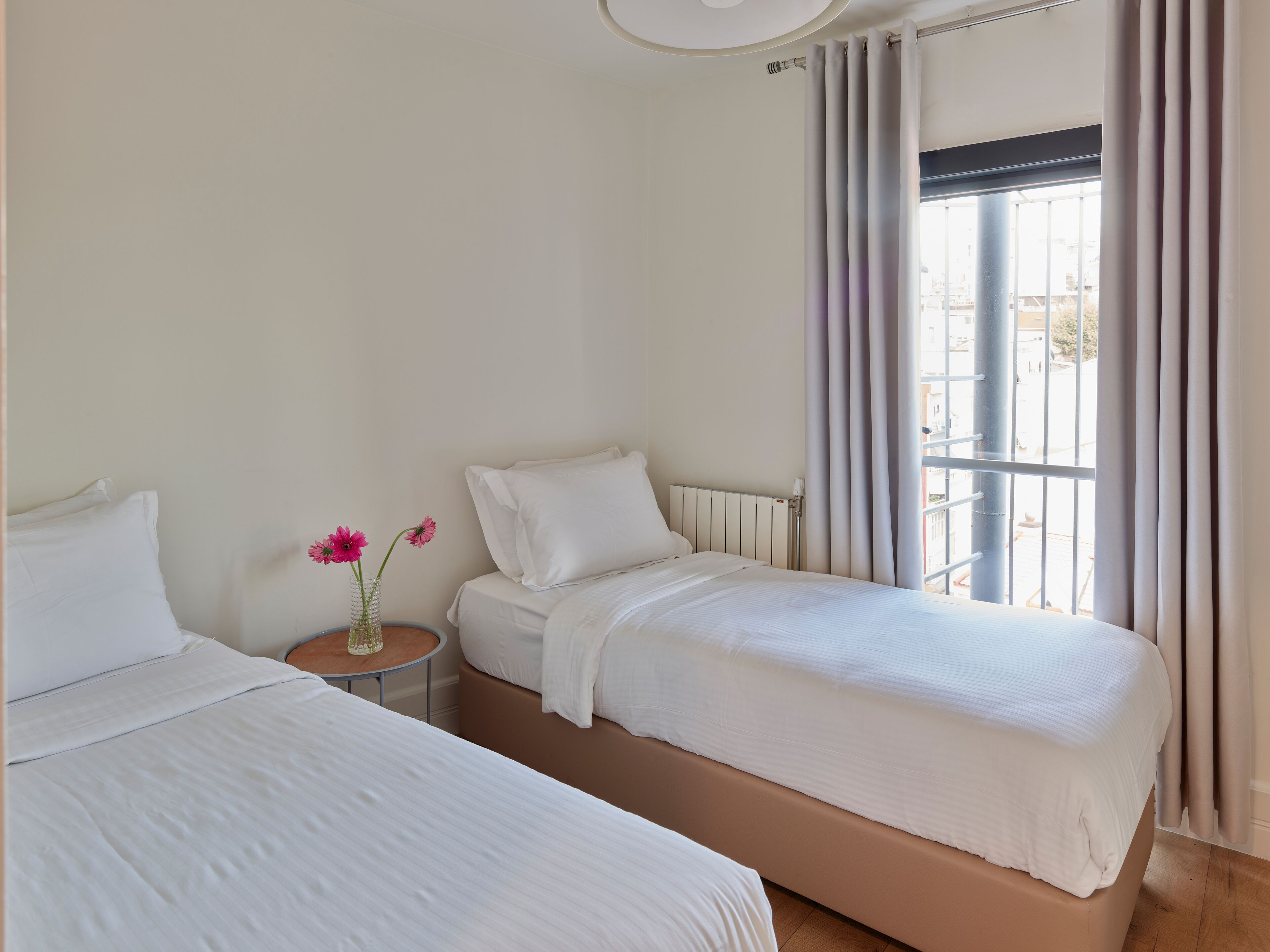 A hotel room with two beds, a small table with a vase of flowers, and a window with curtains.