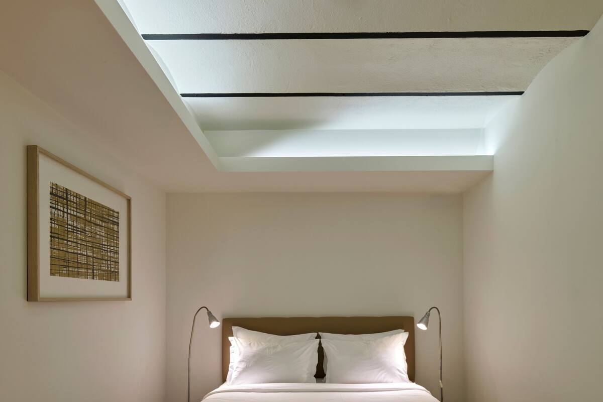 A bedroom with a skylight, a bed with two pillows, a bedside lamp, and a framed artwork on the wall.