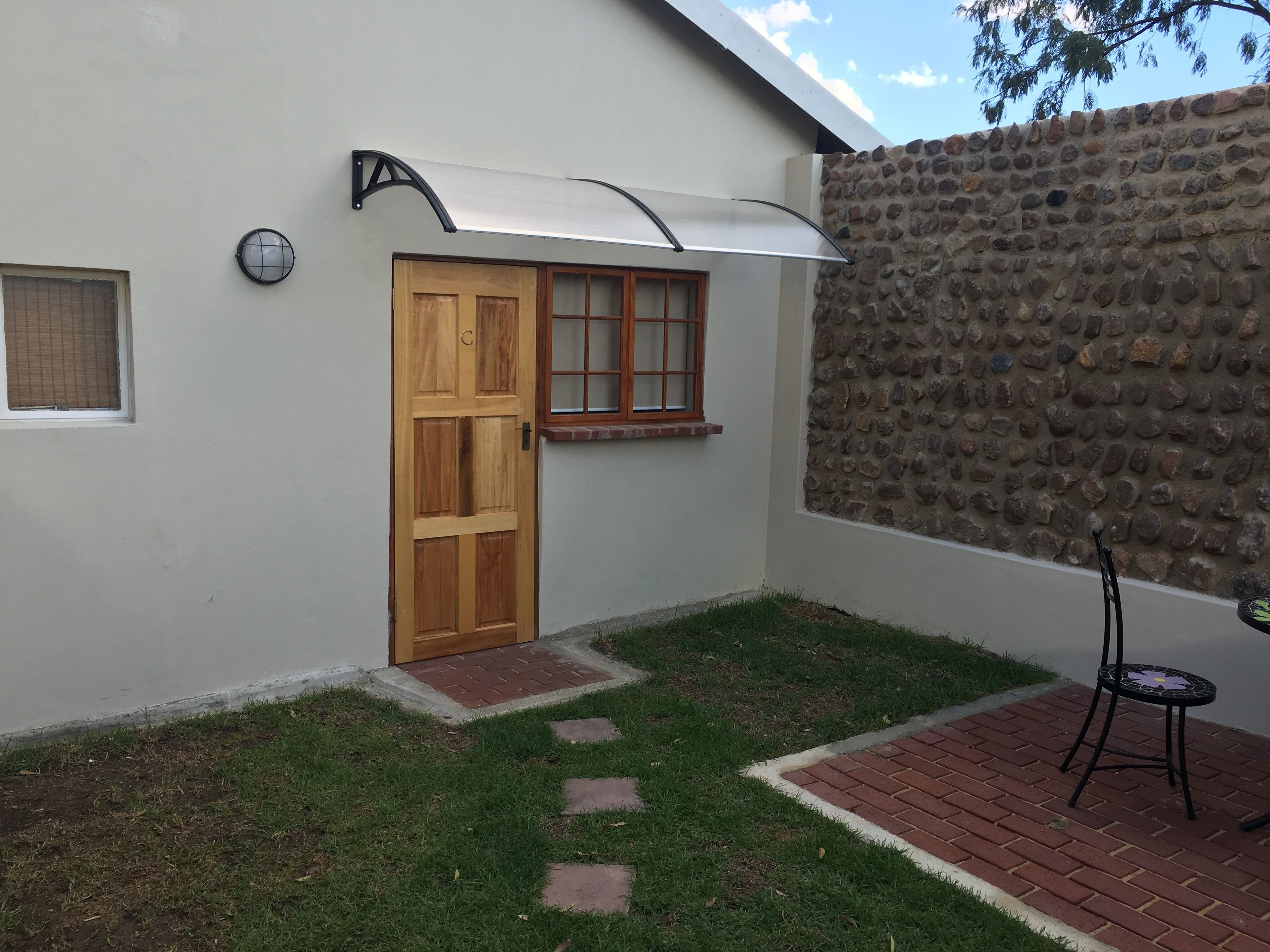 A small house with a wooden door, a window with a grid pattern, a stone wall, and a small table with a chair.