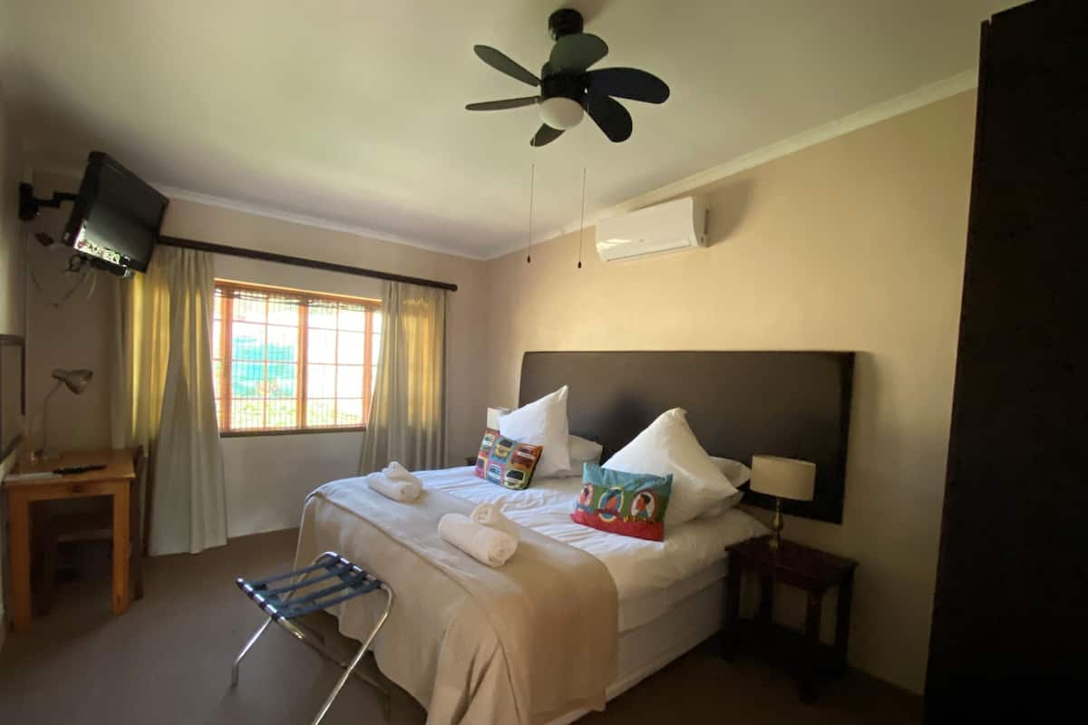 A hotel room with a bed, a ceiling fan, a TV mounted on the wall, a window with curtains, a bedside table, and a chair.
