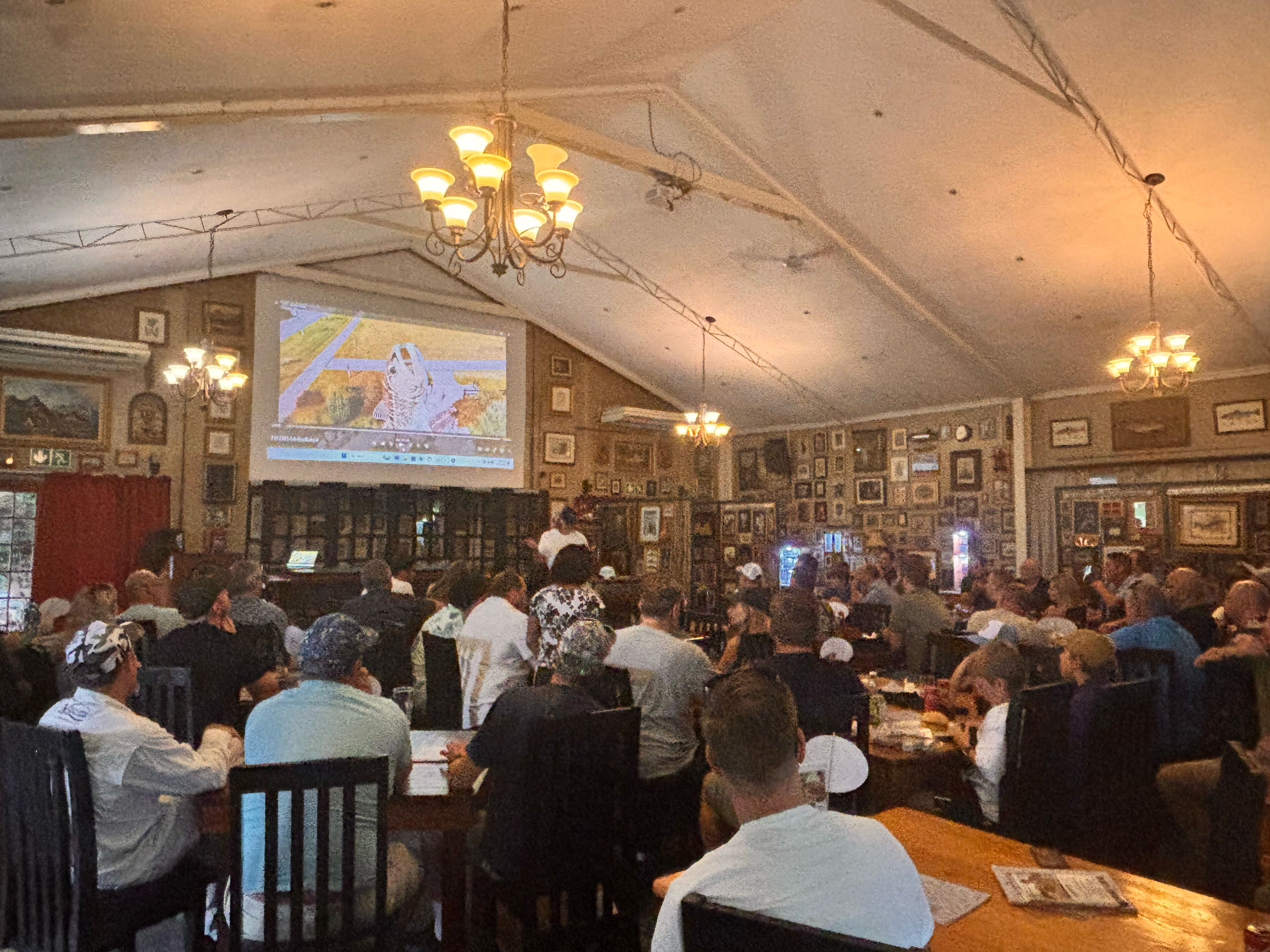 A crowded bar with patrons seated at tables, a large screen displaying a presentation, and multiple chandeliers hanging from the ceiling.