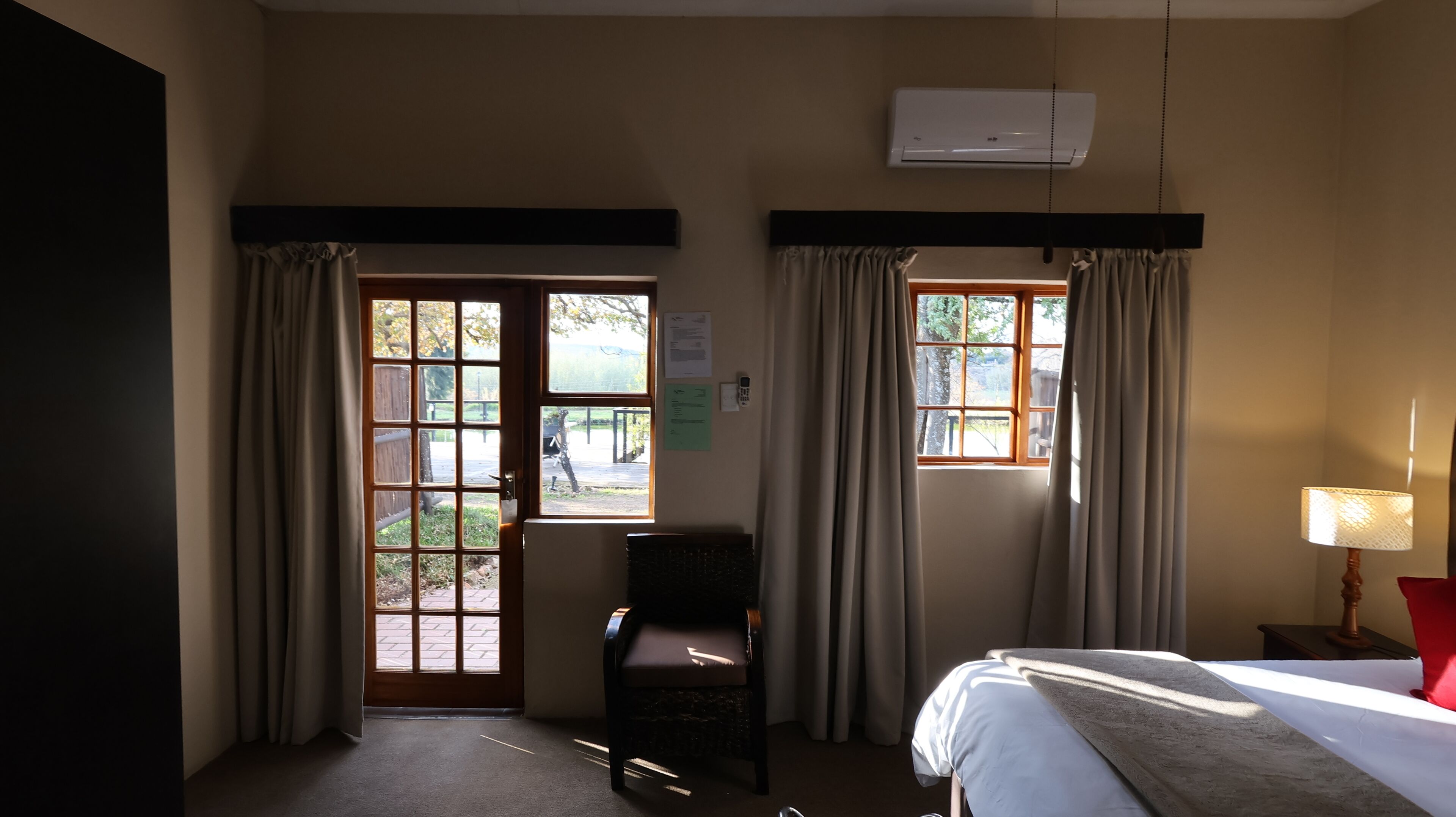 A hotel room with a bed, a chair, two windows with curtains, an air conditioning unit, and a lamp.