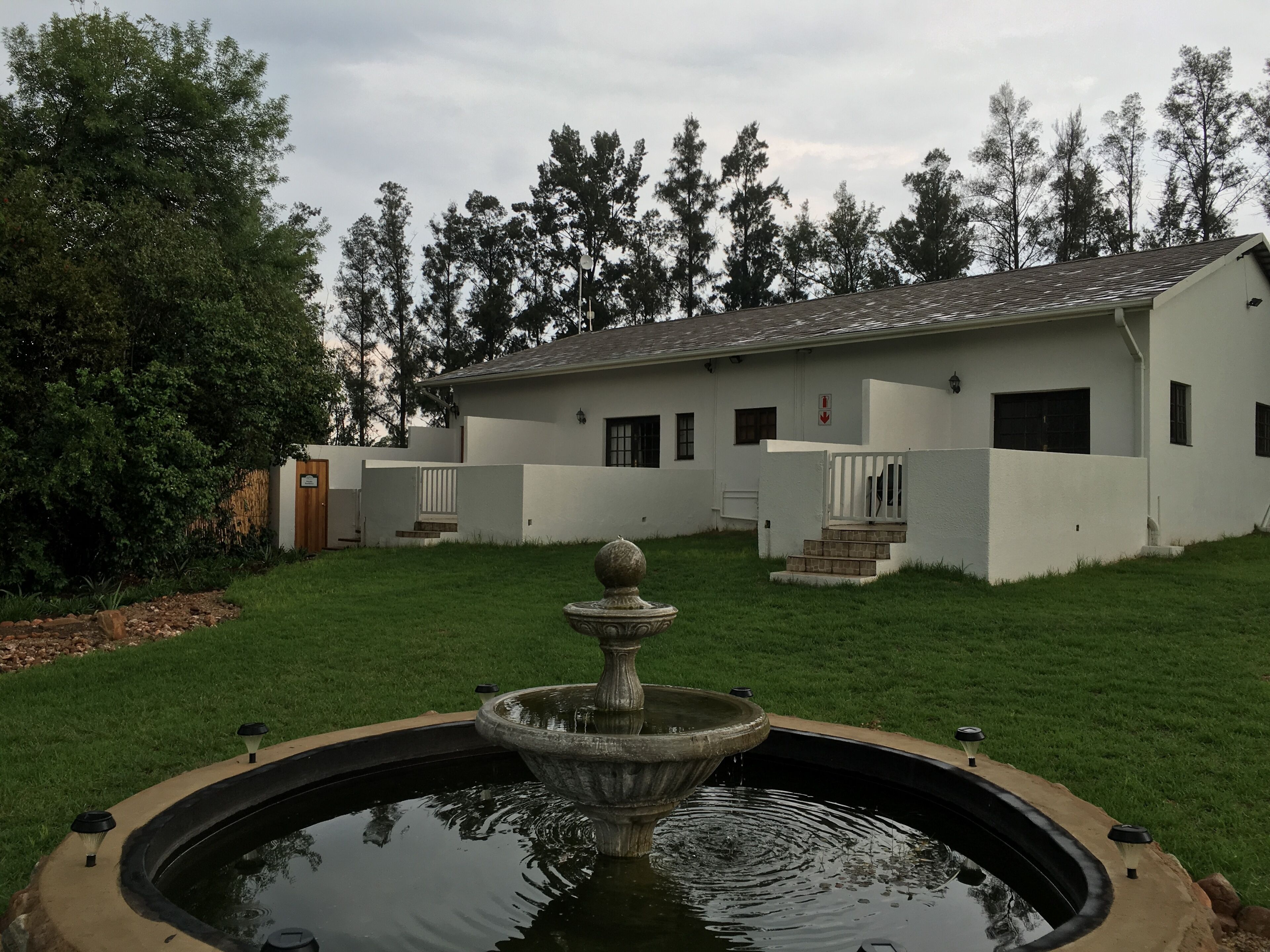 A white building with a fountain in the center, surrounded by a well-maintained lawn and trees.