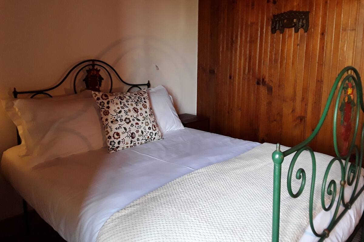 Una cama con almohada floral, ropa blanca y cabecera de metal verde.