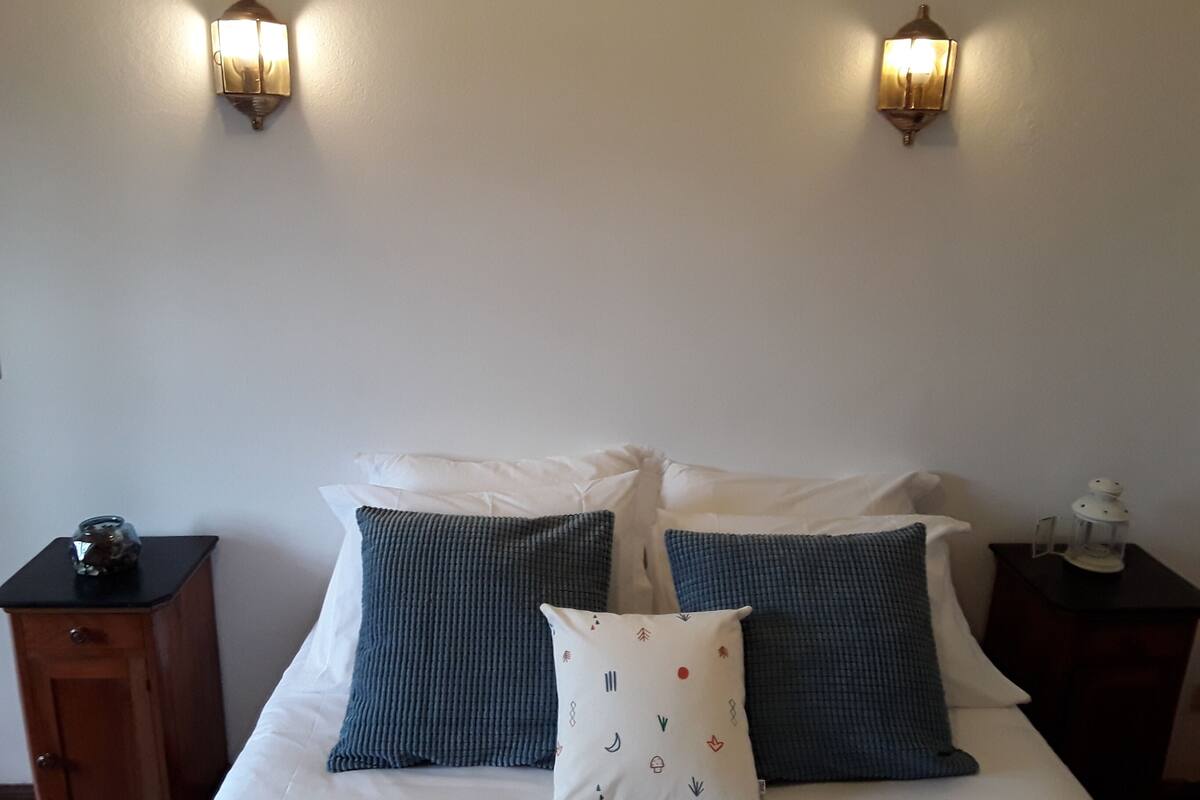 Una cama con ropa de cama blanca y dos almohadas azules, dos lámparas de pared, una mesita de noche de madera con una lámpara y un farol decorativo a la derecha.