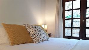 A bed with white bedding and two decorative pillows, a bedside lamp, and a view through a wooden door.