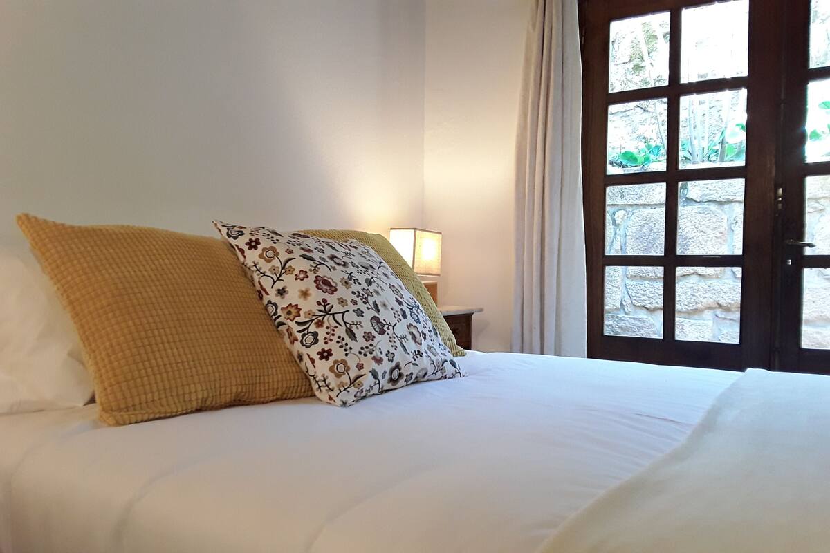 Una cama con ropa blanca y dos almohadones decorativos, una lámpara de noche y vista a través de una puerta de madera.