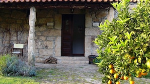 A stone building with a red-tiled roof, a wooden door, and a sign indicating 'Patio A'. There is a pile of firewood and a potted plant in front.
