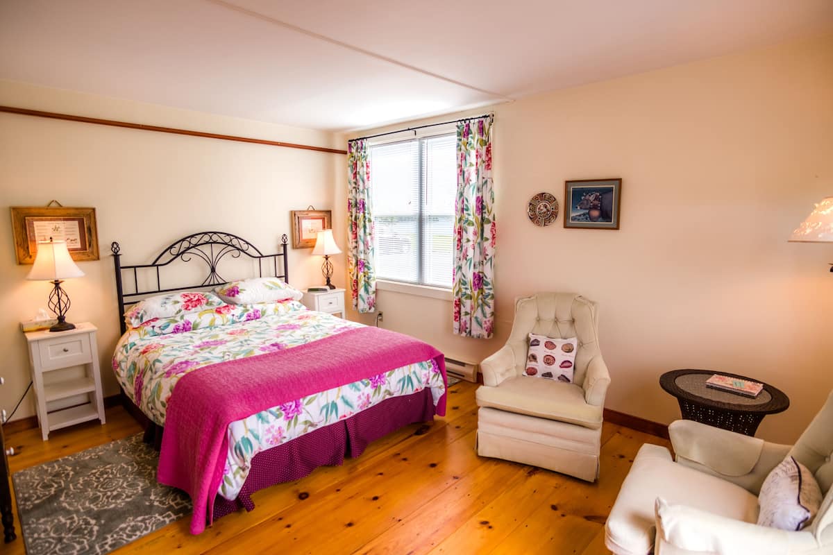 Ein Schlafzimmer mit einem Bett, zwei Nachttischen, einem Sessel, einem kleinen Tisch, einem Fenster mit Vorhängen und Wanddekorationen.
