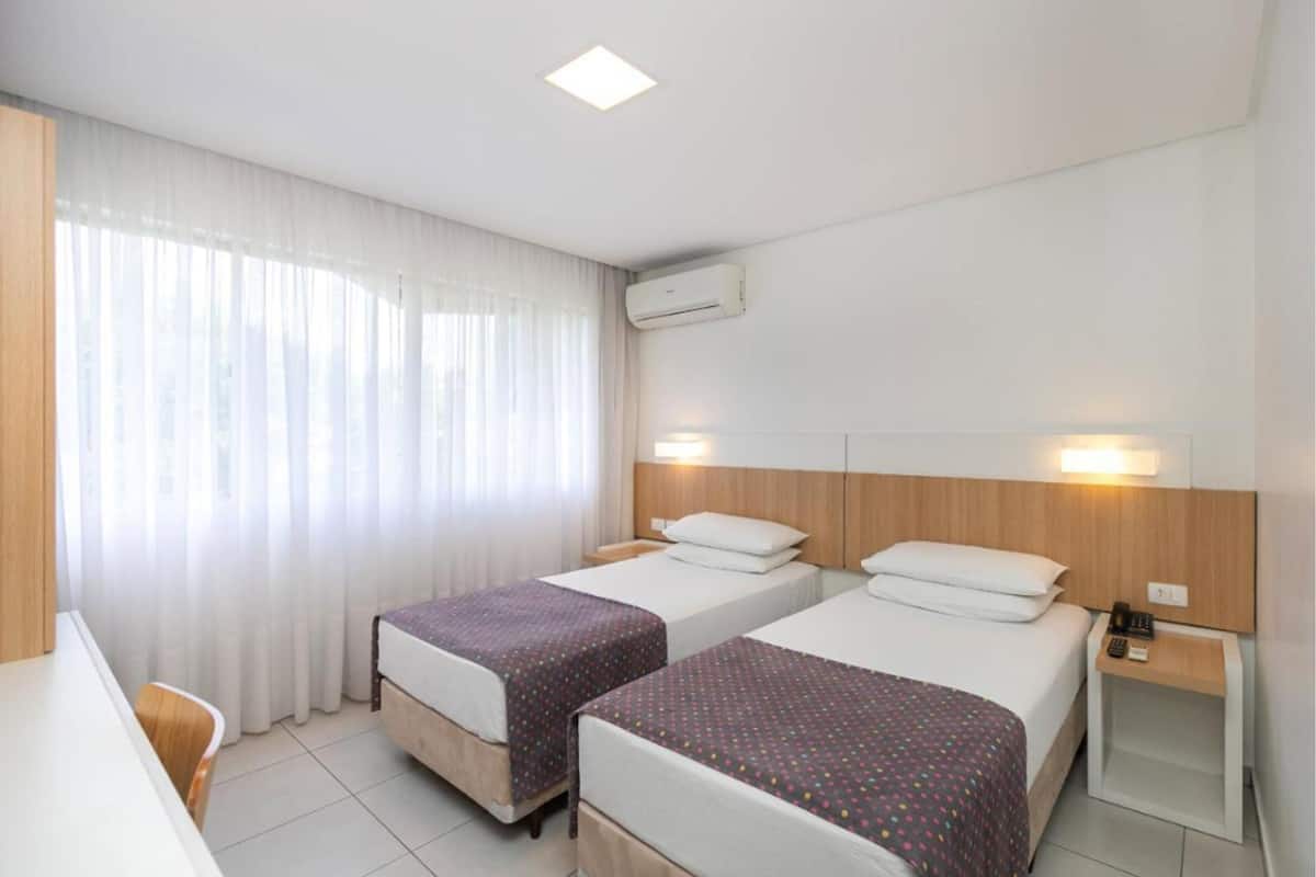 Ein Hotelzimmer mit zwei Einzelbetten, einem Schreibtisch, einem Stuhl und einem Fenster mit leichtem Vorhang.