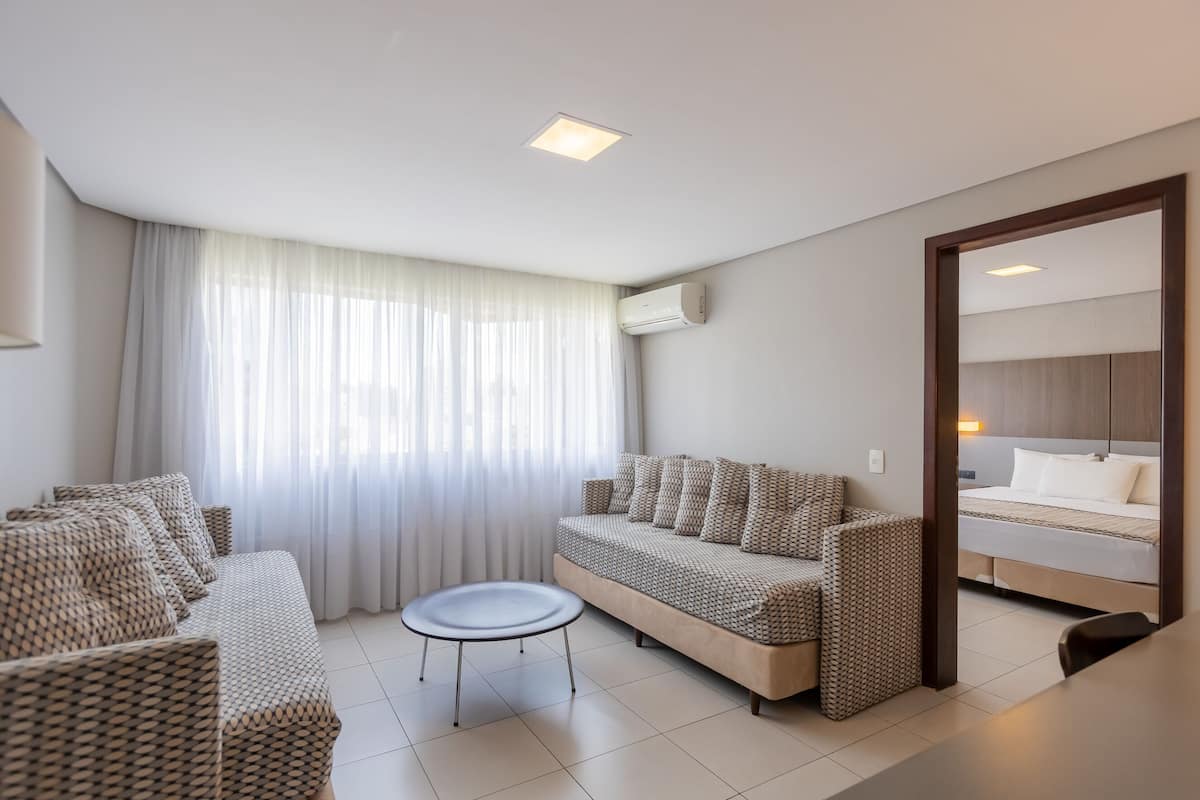 Ein modernes Hotelzimmer mit Sofa, Sessel, rundem Tisch und einem Bett mit Kopfteil.