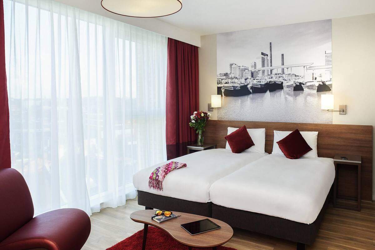 A hotel room with a large bed, a desk, a chair, a lamp, and a cityscape painting on the wall.