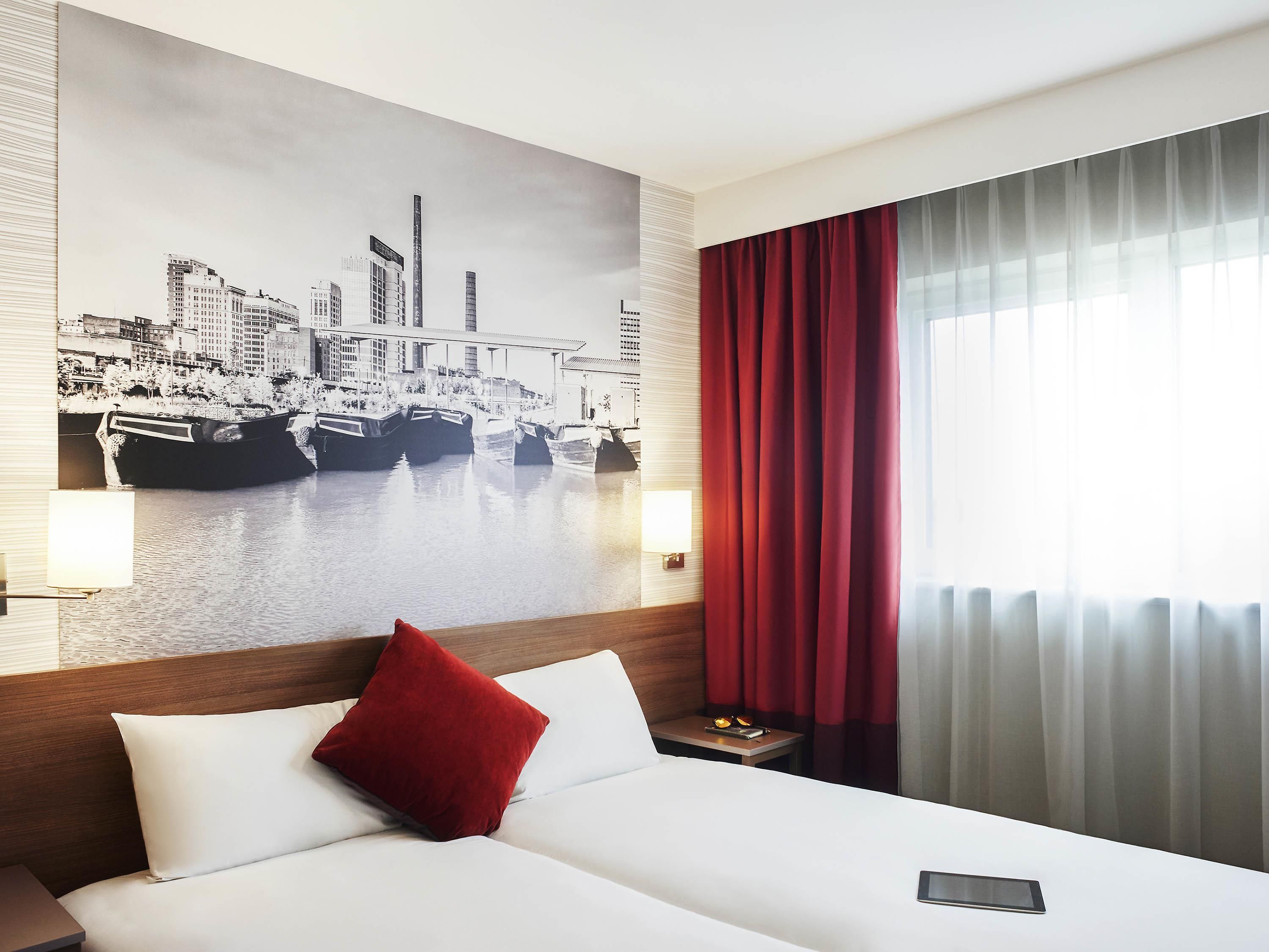 A hotel room with a large bed, a red pillow, a cityscape mural on the wall, a nightstand, and a window with red curtains.