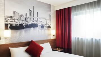 Habitación de hotel con una cama grande, un cojín rojo, un mural de la ciudad en la pared, una mesita de noche y una ventana con cortinas rojas.