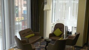 Habitación de hotel con un ventanal grande, un escritorio con documentos y un bolígrafo, dos sillones marrones con cojines verdes y una lámpara de pie.