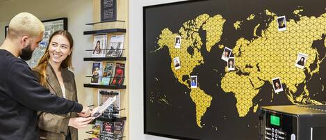 Un hombre y una mujer observan un mostrador con varias revistas, un mapa mundial con fotografías sujetas, y un quiosco digital.