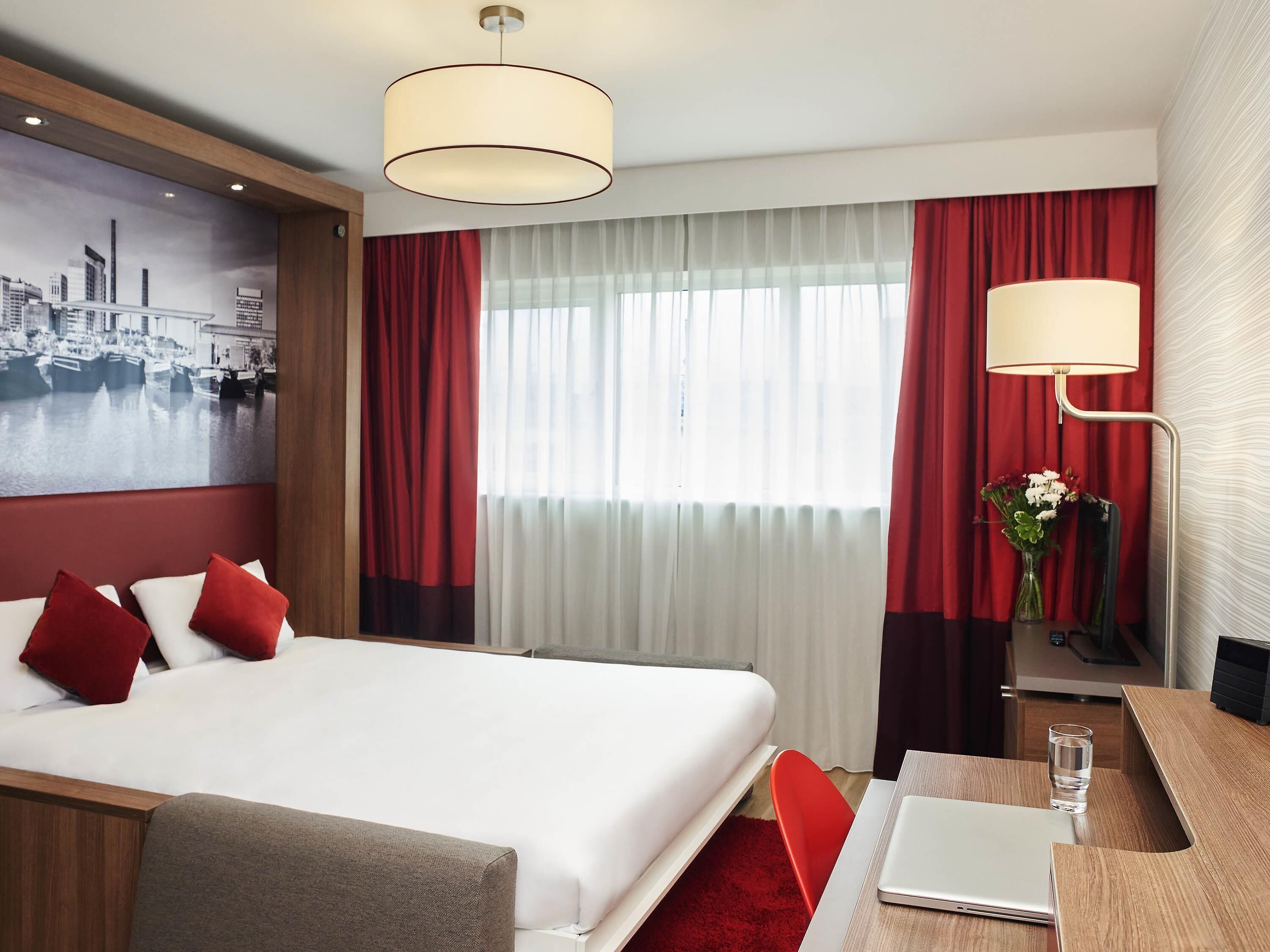 A hotel room with a bed, a desk with a laptop, a vase with flowers, and a cityscape painting on the wall.