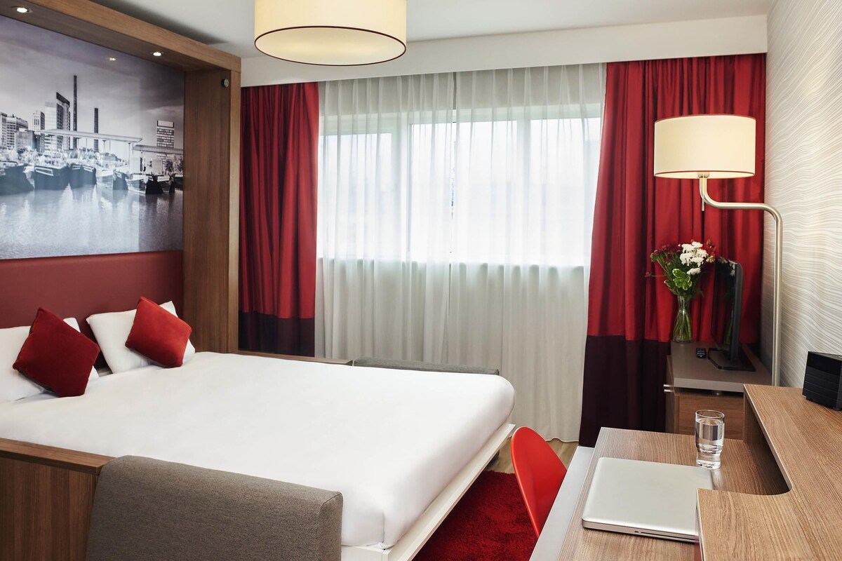 A hotel room with a bed, a desk with a laptop, a vase with flowers, and a cityscape painting on the wall.