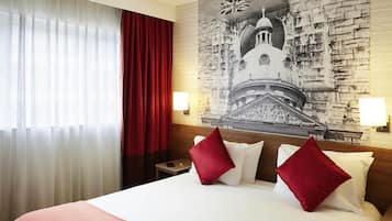 A hotel room with a large bed, red curtains, a wall-mounted lamp, and a mural of a historical building.
