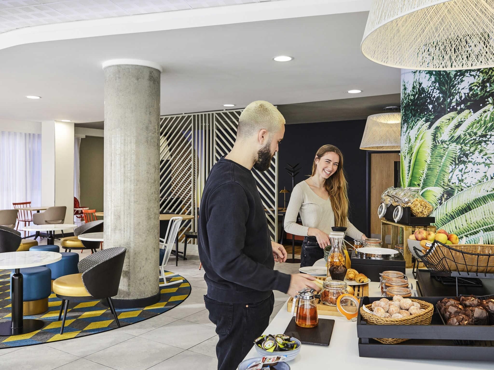 A modern hotel lobby with a central seating area, a buffet with various food items, and a large decorative plant.