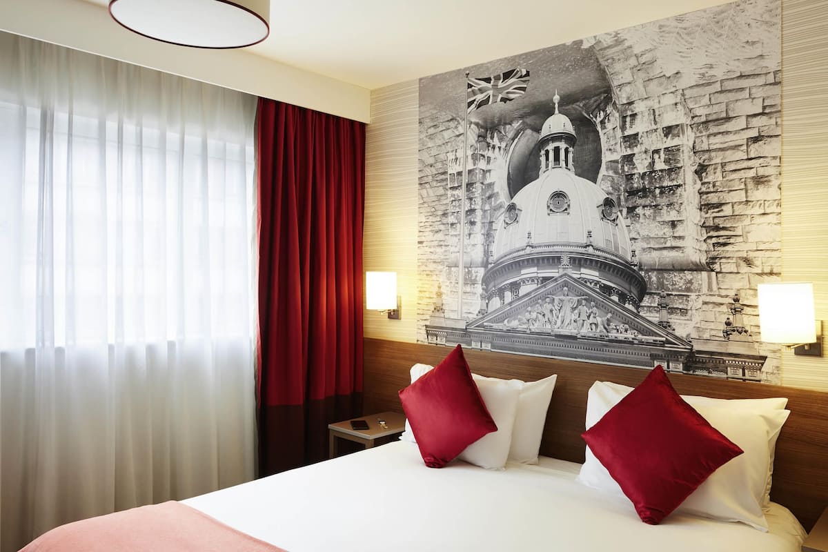A hotel room with a large bed, red curtains, a wall-mounted lamp, and a mural of a historical building.