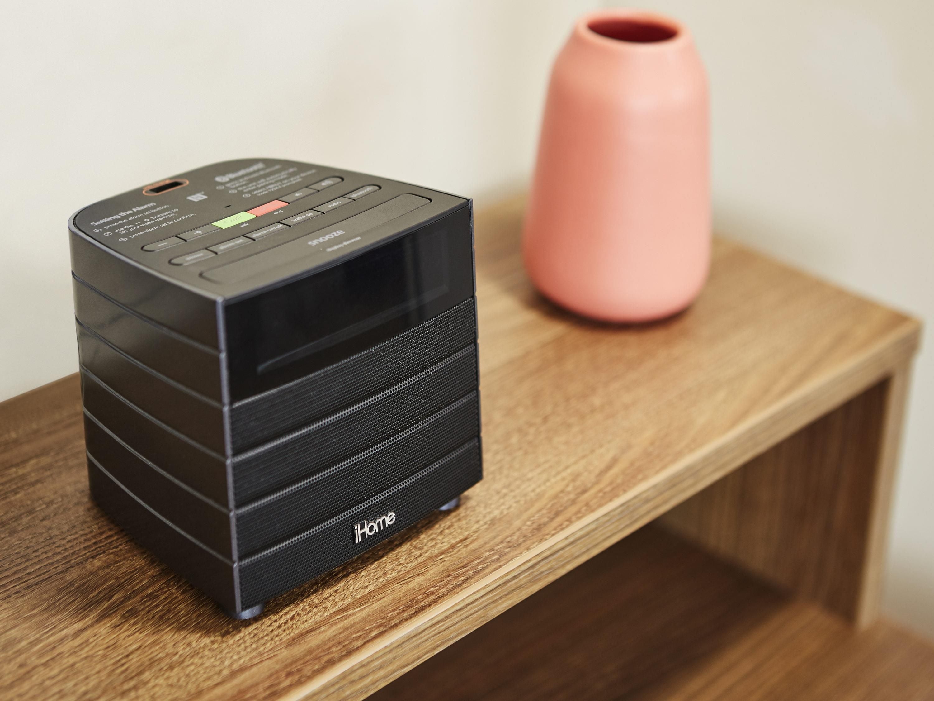 A black iHome speaker on a wooden shelf with a pink vase in the background.