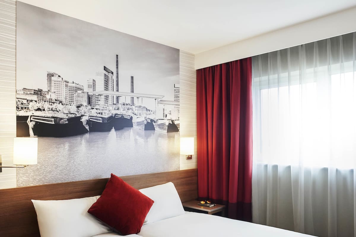 A hotel room with a large bed, a red pillow, a cityscape mural on the wall, a nightstand, and a window with red curtains.