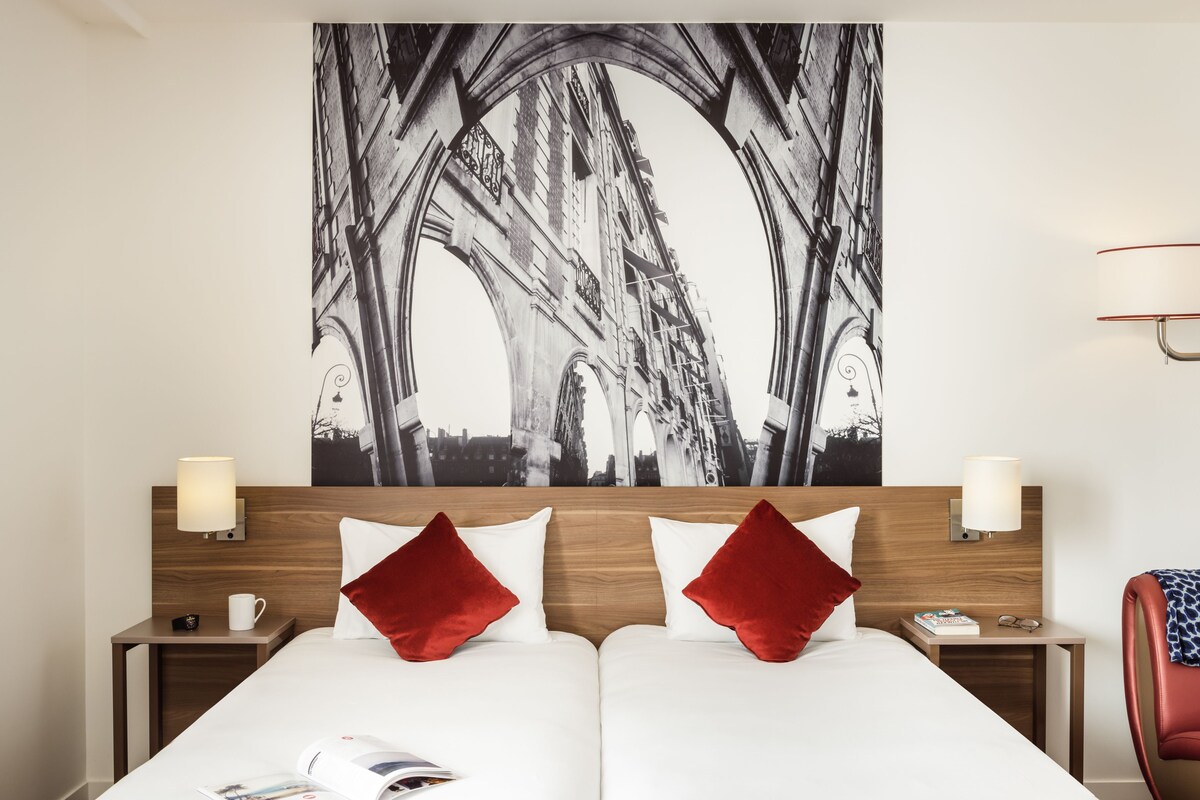 A hotel room with two beds, a large abstract painting on the wall, wooden headboards, bedside lamps, and a red chair.