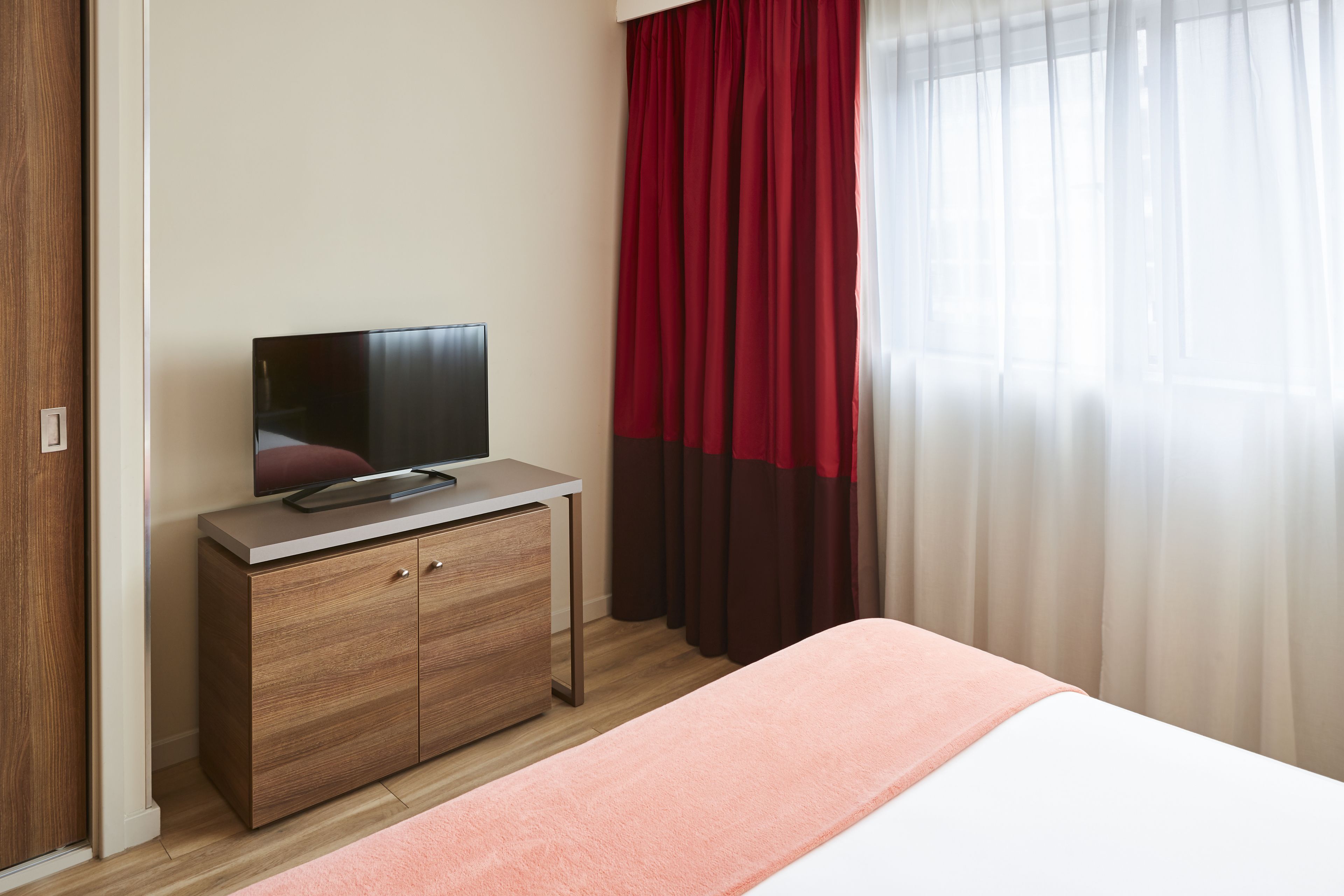A hotel room with a bed, a TV on a wooden cabinet, and red curtains.