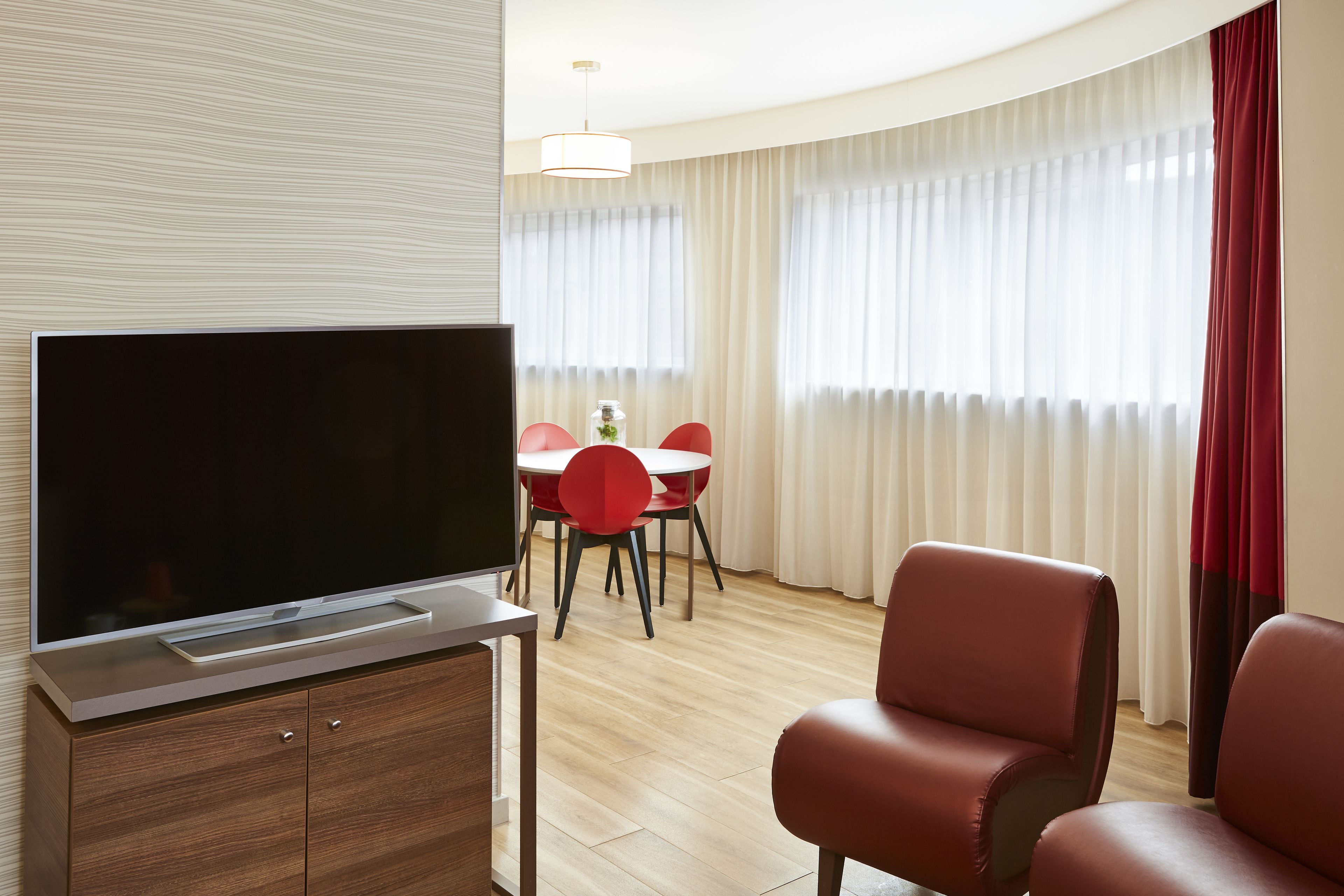 Una habitación de hotel moderna con un televisor de pantalla plana sobre un mueble de madera, una zona de comedor con sillas rojas y un ventanal con cortinas traslúcidas.