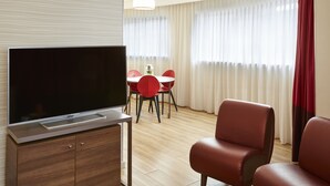 A modern hotel room with a flat-screen TV on a wooden cabinet, a dining area with red chairs, and a large window with sheer curtains. - Aparthotel Adagio Birmingham City Centre (Birmingham)