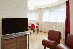 A modern hotel room with a flat-screen TV on a wooden cabinet, a dining area with red chairs, and a large window with sheer curtains. - Aparthotel Adagio Birmingham City Centre (Birmingham)