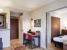 Une chambre d’hôtel compacte comprenant un coin cuisine, un bureau avec une chaise, un lit et une porte donnant sur une autre pièce.