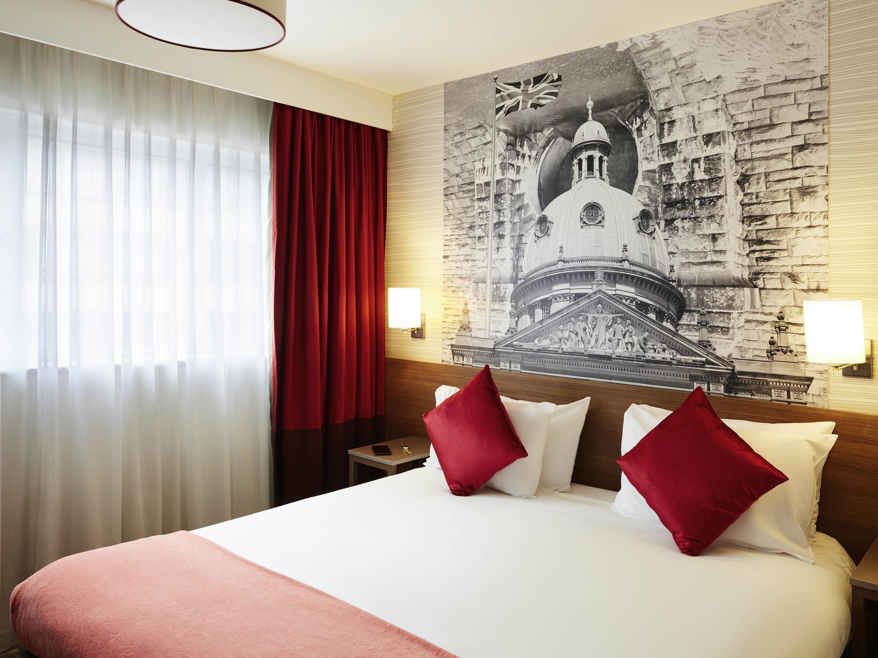 A hotel room with a large bed, red curtains, a wall-mounted lamp, and a mural of a building.