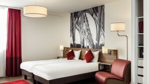 Una habitación de hotel con una cama grande, dos mesitas de noche, una silla roja, una alfombra roja y una obra de arte grande en blanco y negro en la pared.